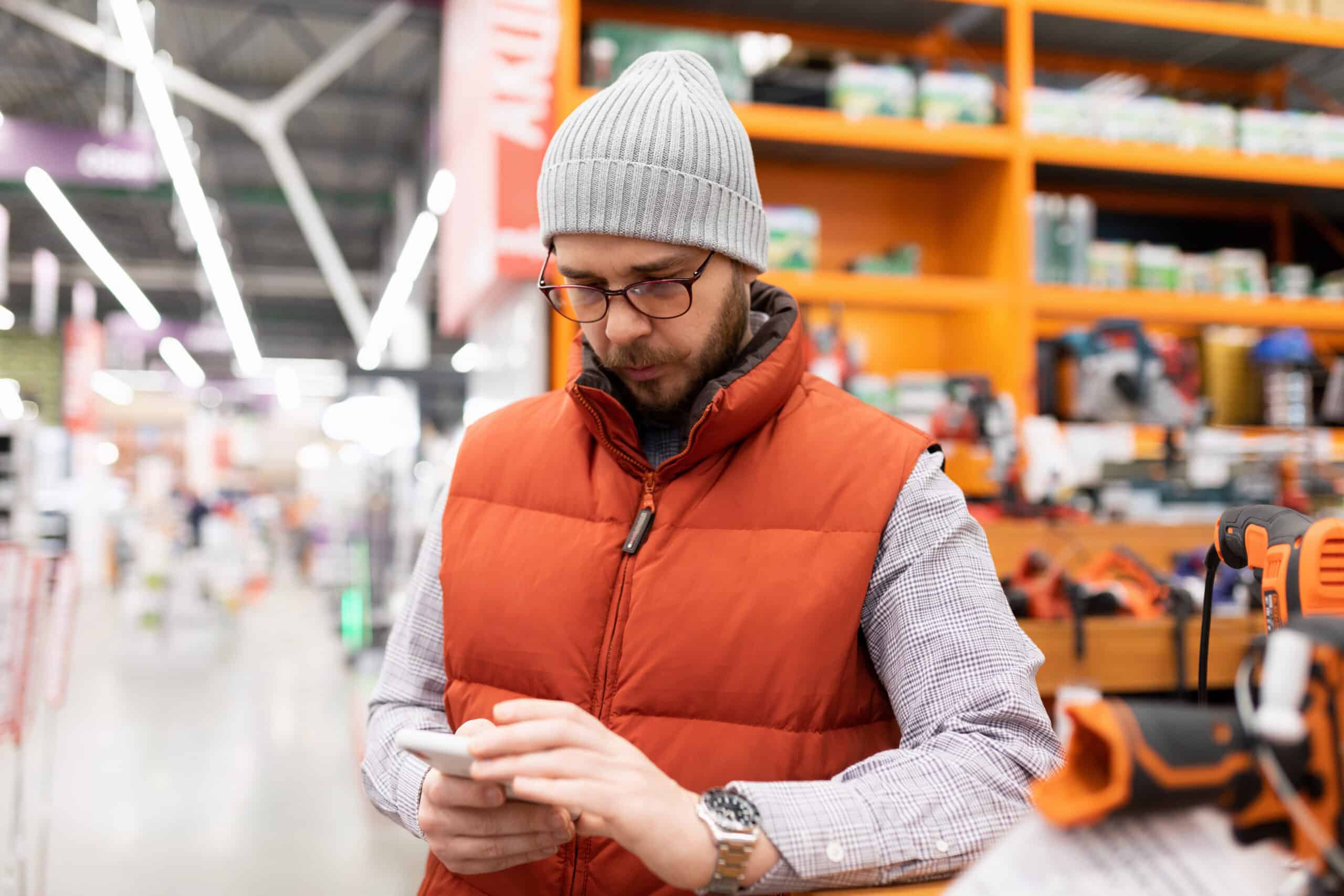 A Man With A Mobile Phone Among The Shelves In A Construction Hypermarket