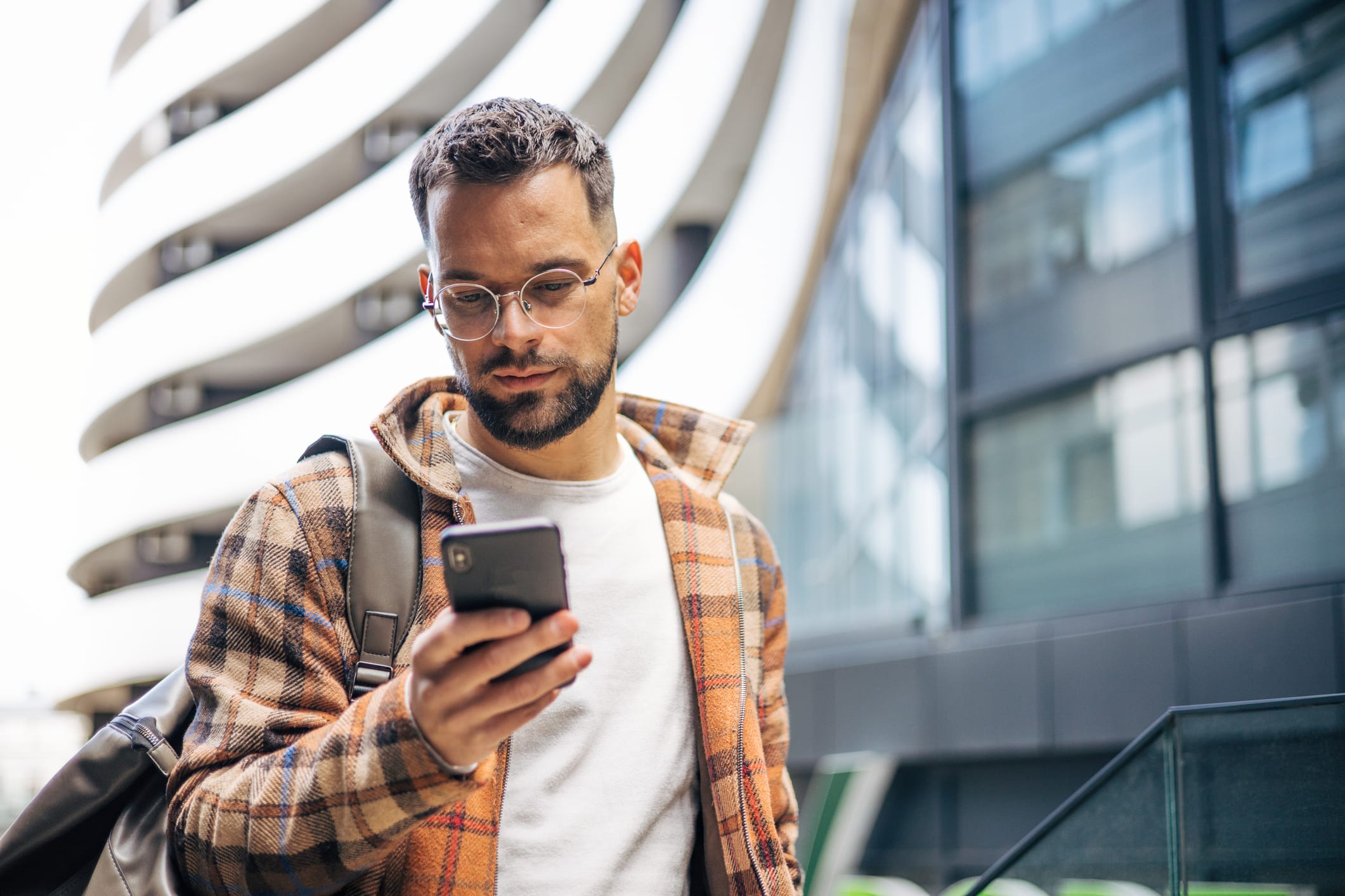 Confused Man Looking Around And Holding His Smartphone. There Are Buildings Around Him.