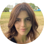 A person with wavy hair is outdoors standing on grass with trees and buildings in the background.