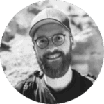 A bearded person smiles wearing glasses and a cap with a blurred outdoor background.
