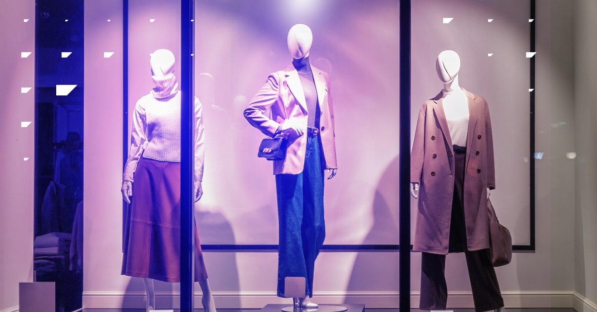 Three mannequins display autumn outfits in a store window each wearing stylish coats and trousers.